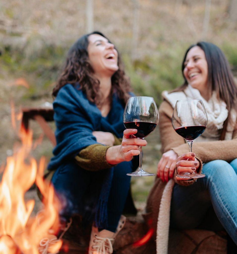 women laughing, holding glass of red wine. Females warming next to the fire.